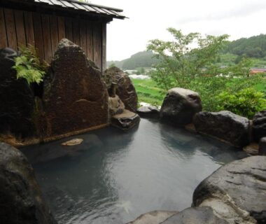 Stone-bordered onsen bath overlooking green hills and wooden wall at Ryokan Yunosako Kumamoto