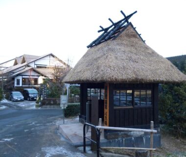 Thatched-roof gate and wooden kiosk beside Ryokan Yunosako with parked cars and courtyard in Kumamoto, Kyūshū