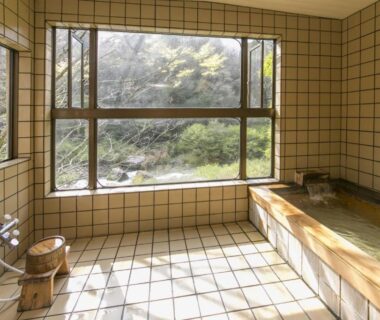 Tiled indoor onsen with wooden soaking tub, stools and large window overlooking a garden in Gifu