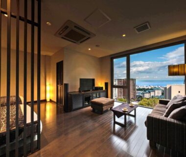 Room with bed, sofa, wooden floor and large window overlooking city and sea at Ryotei Matsubaya Ōita