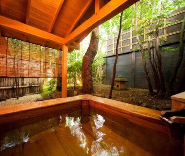 Wooden onsen bath set in a small courtyard with bamboo blinds and trees at Ryotei Tanokura in Ōita