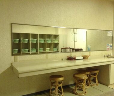 Long mirror and countertop with three woven stools and cubby shelves of green bowls in Ryounkaku wash area