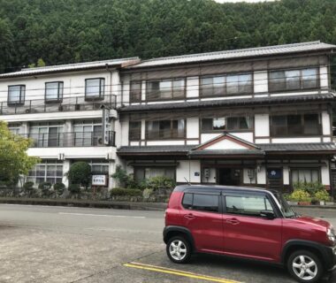 Three-story ryokan facade with balconies, red car in foreground, forested hillside backdrop at Ryujin Onsen Marui Ryokan Wakayama