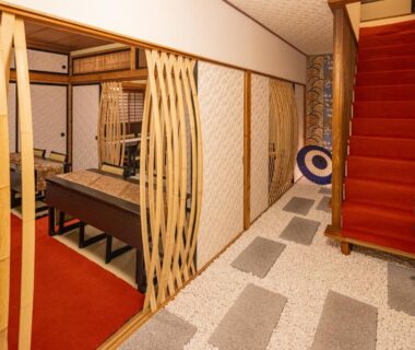 Entrance corridor with wooden slat partitions, low seating area, and red-carpeted staircase in ryokan interior