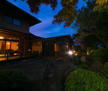 Dusk exterior of wooden ryokan with lit verandah and manicured garden at SEKIYA RESORT Bettei Haruki, Ōita