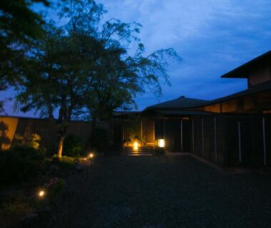 Dusk courtyard with lantern-lit gravel path, trees and wooden ryokan facades at SEKIYA RESORT Bettei Haruki, Ōita