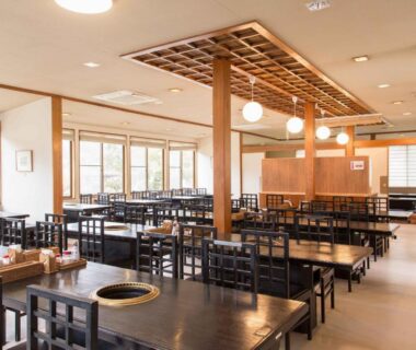 Spacious communal dining hall with wooden tables and chairs, warm lighting, and large lattice windows at Sagirisou Nagano