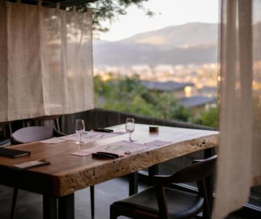 Wooden dining table with glassware and chairs, sheer curtains framing distant city and mountain view