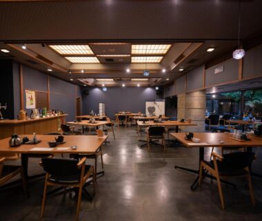 Spacious ryokan dining hall with wooden tables, chairs and warm lighting, large windows overlooking garden greenery