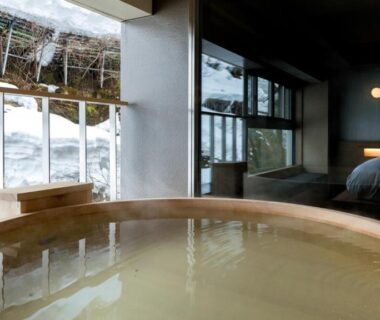 In-room wooden soaking tub beside bed with window overlooking snow-covered garden at Sake Hotel Tamakiya, Niigata