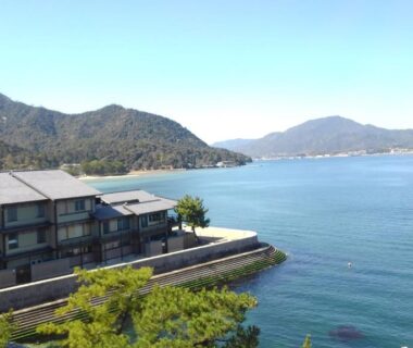 Ryokan buildings on seawall overlooking calm bay with tree-lined shore and distant hills in Hiroshima prefecture
