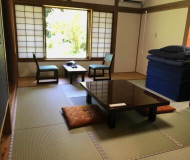 Tatami room with low table, zabuton cushions, stacked futons and shoji window framing green garden view