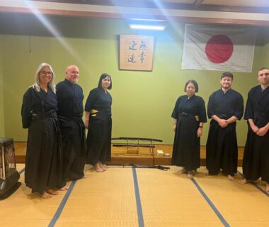 Six guests in traditional attire standing on tatami beneath Japanese flag and calligraphy at Samurai Hotel Naraiso, Nagano