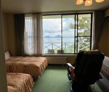 Twin beds with reclining chair and balcony windows overlooking a lake and distant mountains at Marukoma Onsen