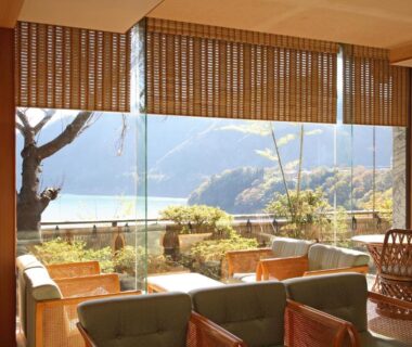 Glass-walled lounge with wicker seating, bamboo blinds and terrace overlooking lakeside and mountains at Sarugakyo Hotel, Gunma