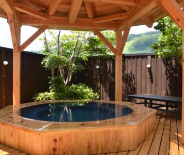 Octagonal wooden outdoor bath under a gazebo on a timber deck with mountain backdrop at Schole Hakuba Nagano