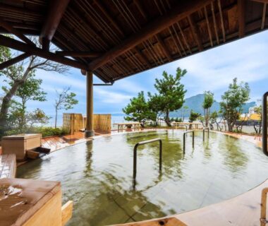 Roofed outdoor onsen pool with handrails, wooden seating, trees and distant coastline at Seaside Hotel Mimatsu Ooetei, Ōita