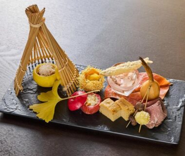 Assorted small Japanese dishes on a black slate plate with bamboo decorative accent and citrus bowl