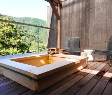 Wooden soaking tub on sunlit deck with chairs and green mountain view at Seikansou, Kanagawa