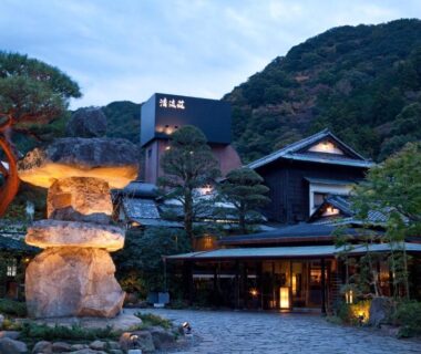 Stone lantern and lantern-lit pathway leading to traditional wooden ryokan entrance with surrounding garden and hillside backdrop in Shizuoka