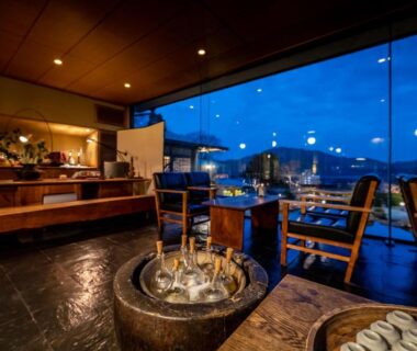 Interior lounge at Sekitei with stone basin, wooden seating and large window overlooking illuminated hills in Hiroshima