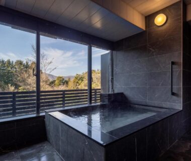 Large stone soaking tub with floor-to-ceiling windows overlooking wooded hillside at Sengokubara COCON, Kanagawa