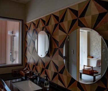 Vanity with twin round mirrors, geometric wood wall, sink and open doorway to guest room at Senkei, Kanagawa