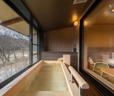 Wooden soaking tub on balcony with bed and seating, large windows framing mountain landscape at Senkyo, Gunma