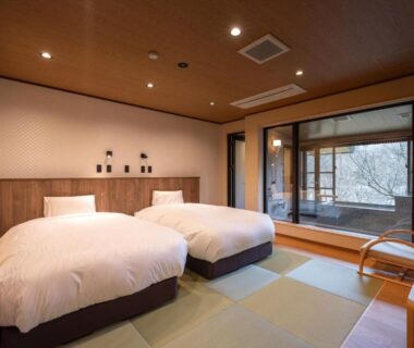 Tatami-floored guest room with twin beds, shoji screens and wooden seating beside large window at Senkyo, Gunma