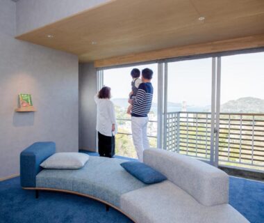Family standing by large sliding window in modern ryokan lounge with curved sofa and balcony overlooking hills