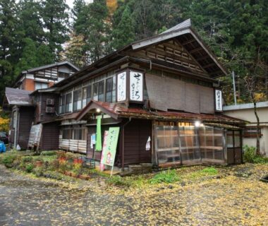 Multi-story wooden ryokan exterior with fallen leaves and entrance banners, forested setting in Niigata, Chūbu, Japan