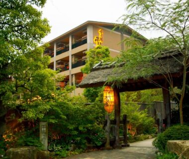 Traditional wooden gate with lanterns and path leading to a multi-story ryokan surrounded by lush trees in Kanagawa