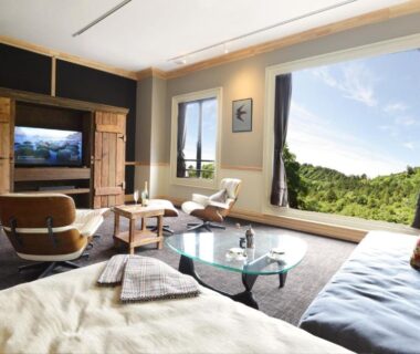Bright ryokan room with wide window overlooking forested hills, low table, sofa and low bed at Shelter Garden Nikko