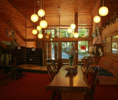 Warm wooden lounge with long dining table, pendant lanterns, seating area and glass doors opening to greenery