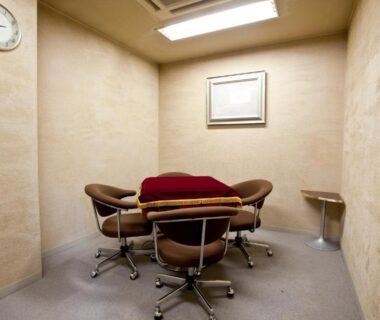 Small beige meeting room with four swivel chairs, low table with red cushion, wall clock and overhead light