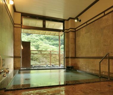 Indoor onsen bath with stone floor, wooden handrail and open doorway to green hillside at Shima Onsen Yoshimoto, Gunma