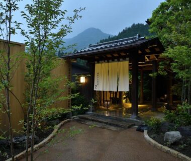 Stone path and wooden gate with noren curtains, lanterns, and surrounding trees at Shima Onsen Yoshimoto, Gunma