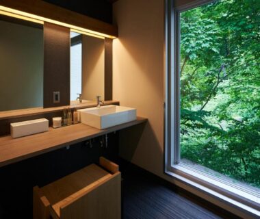 Bathroom vanity and wooden stool overlooking lush forest through large window at Shima Onsen Yuzuriha, Gunma, Kantō