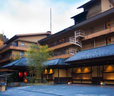 Multi-story wooden ryokan facade with lit entrance, paper lanterns and autumn foliage against a mountain backdrop in Gunma