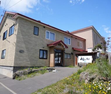 Two-story beige ryokan with red roof, entrance sign and wildflowers along paved path in Hokkaidō