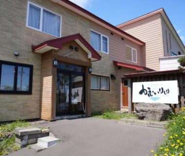 Exterior entrance of Shimanoyado Kamuirishiri with glass doors, signboard, stone feature and small flower beds in Hokkaidō