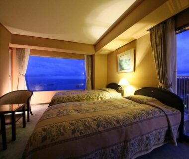 Twin beds, night lamps, seating area, balcony door and sea visible through wide window at Shimoda View Hotel
