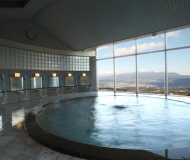 Circular indoor onsen bath, floor-to-ceiling windows framing distant mountains, washing stations along tiled wall at Shinki Gunma