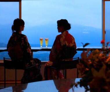 Two guests in yukata by a large window with drinks and mountain view at Shinki ryokan, Gunma, Kantō, Japan