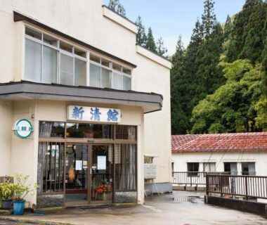Front entrance of Shinseikan with glass doors and signboard, building beside forested hillside in Iwate, Tōhoku