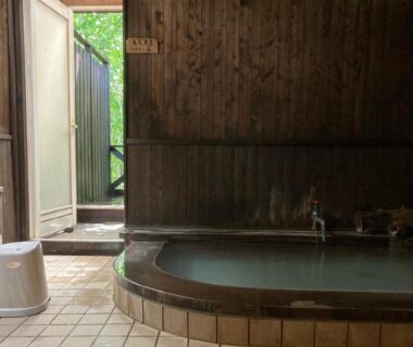 Wooden indoor onsen bath with tiled floor and open doorway to greenery at Shinyu Onsen Kurikomaso, Miyagi