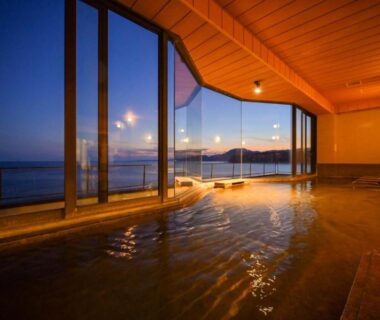 Indoor onsen bath with floor-to-ceiling windows overlooking coastal waters at dusk