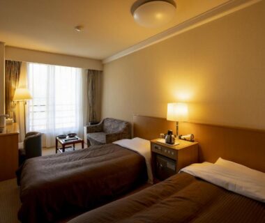 Compact twin guest room with two single beds, bedside lamps, seating area and window at Shirafune Grand Hotel Nagano