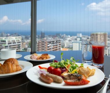 Western-style breakfast plate with croissant, sausages, salad and juice on table by large window overlooking cityscape in Ōita