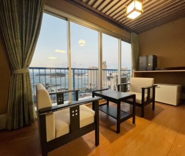 Room with sliding glass doors leading to balcony, two armchairs and low table overlooking distant water and city